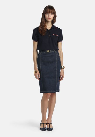 Woman standing wearing a navy short-sleeve polo, high-waist dark denim pencil skirt, black pointed-toe T-strap heels, and gold accessories.