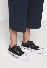 Black canvas sneakers with white rubber sole and accents, featuring eyelets and laces. The shoes are worn with dark cropped pants.