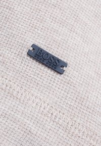 Beige texturerad tyg med en svart rektangulär etikett sydd på, präglad med "BOSS HUGO BOSS"-märke.