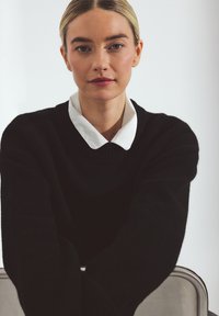 Black sweater with a ribbed neckline layered over a white collared shirt, seated against a plain background. Smooth texture and simple design.