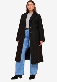 Lang sort uldfrakke med klippet krave, to forreste knapper og sidelommer, parret med lyseblå vidde jeans og sorte ankelstøvler.