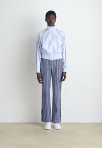 Woman wearing a white zip-up jacket with patterned blue pants and white sneakers standing against a plain white wall.