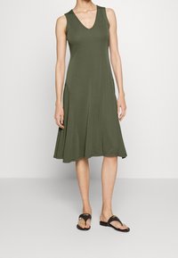 Modelka ubrana w bezrękawnikową, oliwkową sukienkę z dekoltem w kształcie V, w połączeniu z czarnymi klapkami, stoi na jednolitym tle.