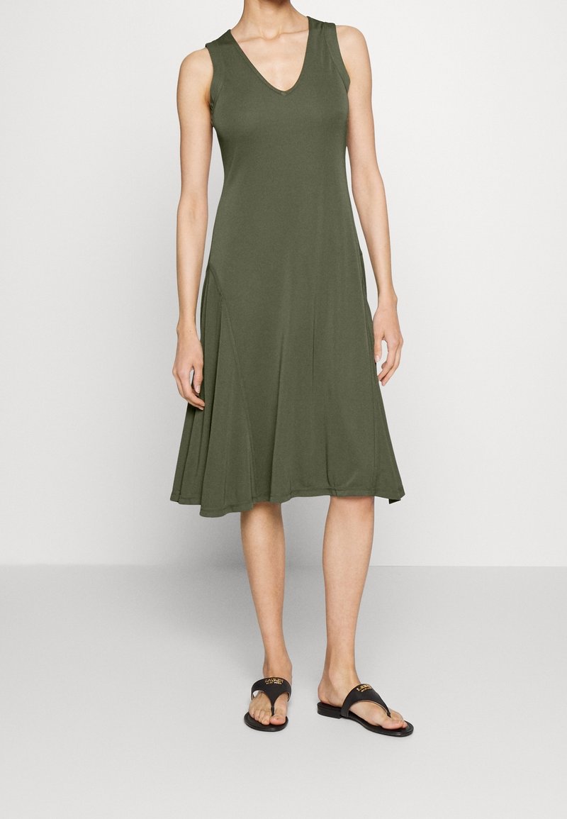 Modelka ubrana w bezrękawnikową, oliwkową sukienkę z dekoltem w kształcie V, w połączeniu z czarnymi klapkami, stoi na jednolitym tle.