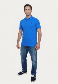 Ung man står klädd i en blå polo, blå jeans och svarta sneakers mot en enkel ljus bakgrund.