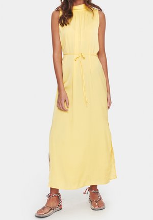 Vrouw die een mouwloze gele maxi-jurk draagt met een ceintuur in de taille en sandalen met meerkleurige gevlochten bandjes, staand tegen een witte achtergrond.