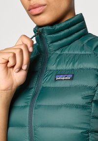 Jakna s punjenjem u tamnozelenoj boji, s horizontalnim šavovima, visokim ovratnikom i patentnim zatvaračem sprijeda s logotipom Patagonia.
