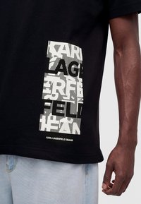 Μαύρο T-shirt με λευκό παραμορφωμένο κάθετο κείμενο και "Karl Lagerfeld Jeans" τυπωμένο στο κάτω μέρος, φορεμένο με ανοιχτό μπλε τζιν.