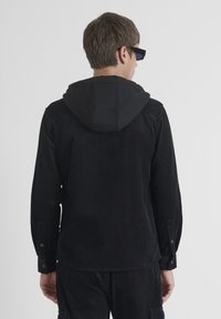 Chaqueta de pana negra con capucha, con puños abotonados y textura de cordones verticales, vista desde la parte posterior, combinada con pantalones a juego.