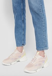 Persona che indossa sneakers chunky rosa chiaro e bianche con jeans di denim su uno sfondo grigio chiaro uniforme.