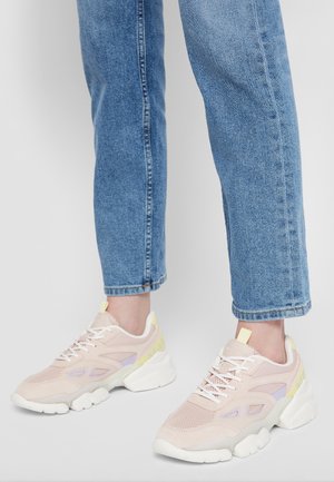 Personne portant des baskets chunky rose clair et blanches avec un jean en denim devant un fond gris clair uni.