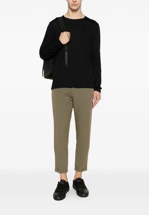 Uomo che indossa una maglia nera a maniche lunghe, pantaloni corti color oliva, sneakers nere e porta uno zaino nero su una spalla.