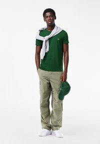 Vihreä lyhythihainen paita, harmaa huppari hartioilla, khaki-väritetyt cargo-housut, valkoiset lenkkarit ja kädessä pidettävä vihreä lippis.