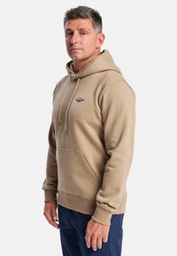Beige hoodie met een voorkeurszak, trekkoord en een klein logo-embleem. Zachte stof met ribgebreide boorden en onderrand. Gekocht over een marineblauwe broek.