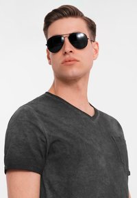 Černé tričko s výstřihem do V a texturovaným povrchem, krátké rukávy, s náprsní kapsou. Model nosí černé sluneční brýle.