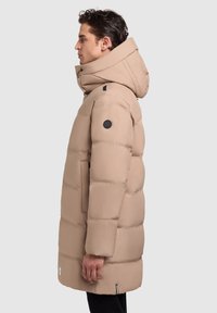 Hombre con abrigo invernal largo y acolchado de color beige, con cuello alto y capucha, de perfil contra un fondo liso.