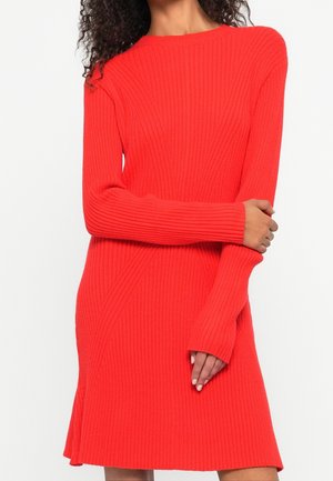 Femme portant une robe en maille côtelée rouge vif à manches longues, debout avec un bras croisé et l'autre main touchant l'ourlet de la robe.