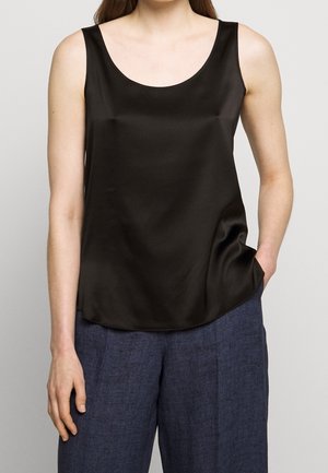 Top negro sin mangas de satén combinado con pantalones anchos azul texturizados, con una mano en el bolsillo y fondo neutro.