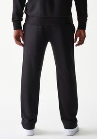 Μαύρο παντελόνι φόρμας από βαμβακερό μείγμα υφάσματος, με άνετη εφαρμογή, πλαϊνές τσέπες και ριμπ μανσέτες. Λευκά αθλητικά παπούτσια φαίνονται στα πόδια.