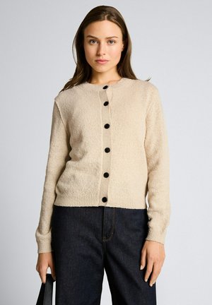 Kobieta ubrana w beżowy sweter na guziki i ciemne dżinsy, trzymająca czarną torebkę, stoi przodem na jednolitym tle.