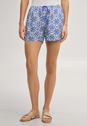 Femme portant un short bleu et blanc à motifs avec une taille à cordon et des sandales beiges à enfiler, debout devant un fond clair.
