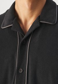 Musta kauluspaita, jossa on teksturoitu kangas, harmailla kontrastiväreillä varustettu piping ja musta nappilistat. Lähikuva kauluksesta ja napeista.