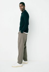 Black long-sleeve polo shirt, beige trousers, and white sneakers. Model stands sideways against a plain background, hands in pockets.
