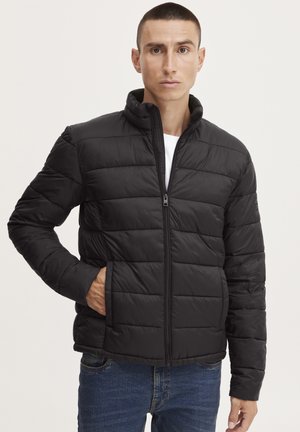 Sort polstret jakke med lynlås foran, stående krave og vandret syninger. Båret over en hvid skjorte med casual blå jeans.