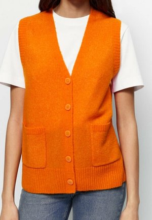 Woman wearing a bright orange buttoned knit vest with front pockets over a white short-sleeve shirt and blue jeans.