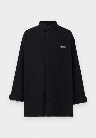 Camisa negra holgada con cuello, que cuenta con dos bolsillos frontales y puños deshilachados. Está hecha de una tela resistente con un sutil logotipo en el pecho.