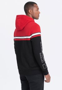 Černá a červená mikina s kapucí, s bílými pruhy přes hrudník a šedým textem na rukávu. Měkká tkanina, sportovní design.