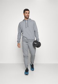 Hoodie de sport gris avec une poche kangourou et un cordon de serrage, associé à un pantalon de survêtement assorti. Plaques de poids noires en main, baskets bleues.
