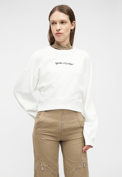 Femme portant un sweat-shirt blanc avec un texte en écriture cursive et un pantalon beige taille haute avec des zips à l'avant et aux genoux, debout devant un fond clair.