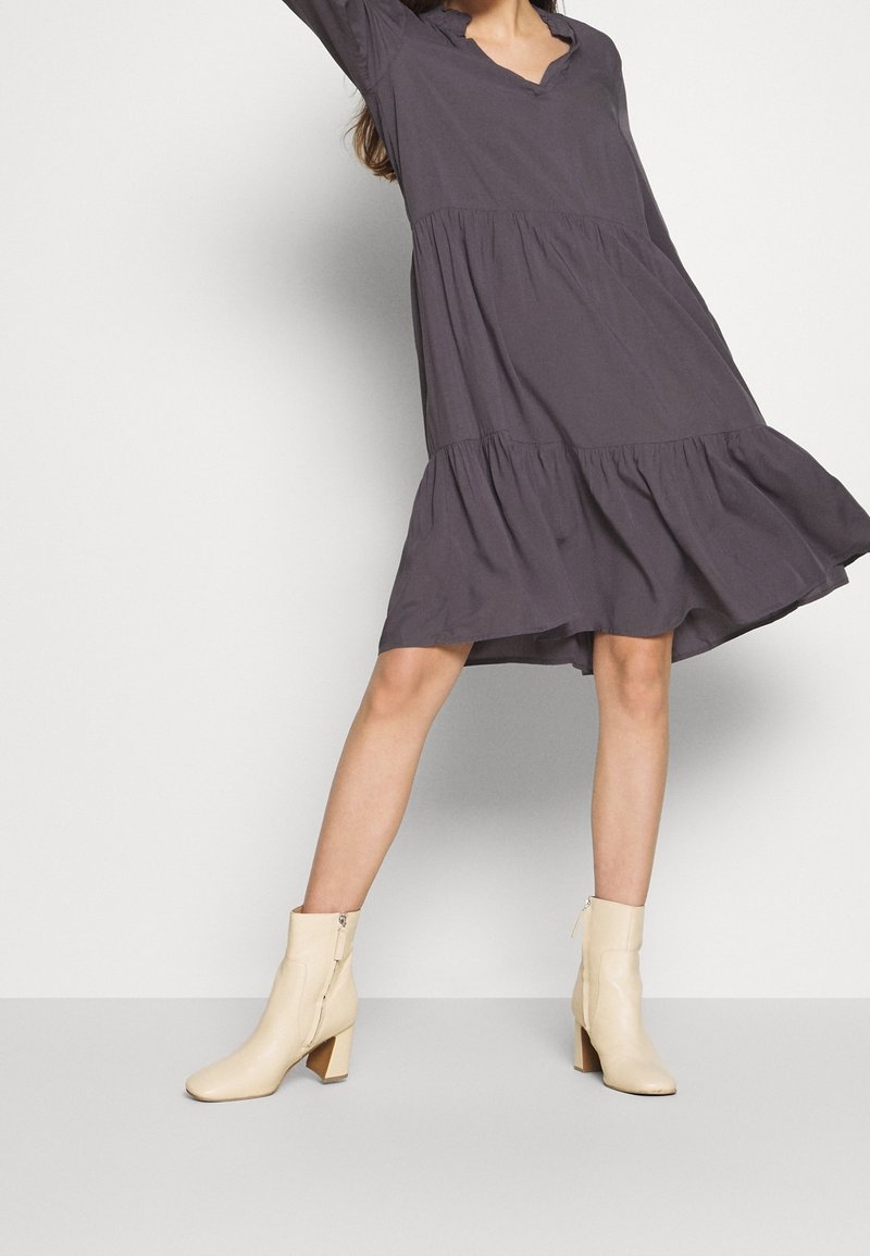 Femme portant une robe à volants gris foncé et des bottines beige clair, posant avec un bras levé sur un fond uni.
