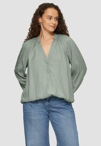 Grüne Bluse mit langen Ärmeln aus leichtem Stoff, mit V-Ausschnitt und gerafften Details an den Schultern, kombiniert mit hellblauen Jeans.