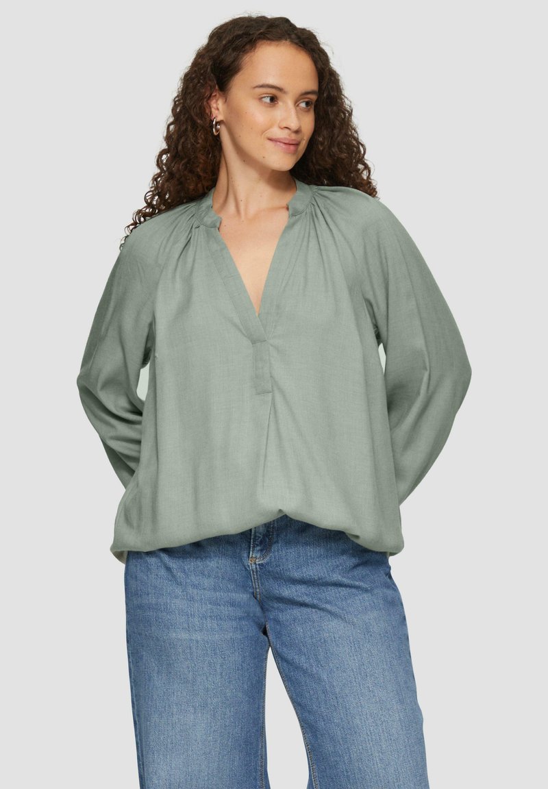 Grüne Bluse mit langen Ärmeln aus leichtem Stoff, mit V-Ausschnitt und gerafften Details an den Schultern, kombiniert mit hellblauen Jeans.