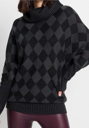 Femme portant un pull à col roulé à motif losanges gris foncé et noir, leggings en cuir bordeaux, main dans la poche.