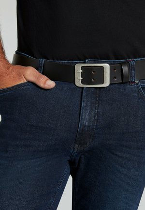 Sort læderbælte med en sølv rektangulær spænde, båret over mørkeblå denim jeans. Spændet har en børstet finish og synlige huller.