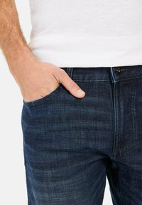 Jean en denim bleu foncé avec une coupe slim, doté d'une poche avant et d'un léger délavage. Le tissu présente une texture lisse et un design classique.