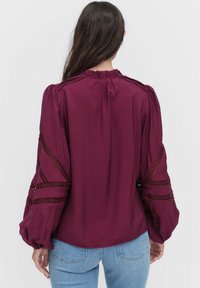 Frau mit langen braunen Haaren, die eine burgunderfarbene Bluse mit Spitzenakzenten an den puffigen Ärmeln und hellblauen Jeans trägt, steht mit dem Rücken zur Kamera.