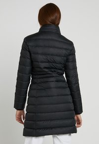 Abrigo acolchado negro con silueta ceñida, patrón acolchado horizontal, cuello alto y mangas largas. Textura de tela lisa y ligeramente brillante.