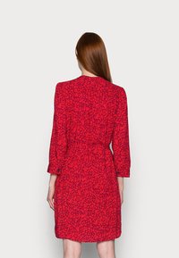 Robe rouge à fleurs avec des manches longues, des poignets à boutons et une taille cintrée. Présente un tissu texturé avec un petit motif florale.