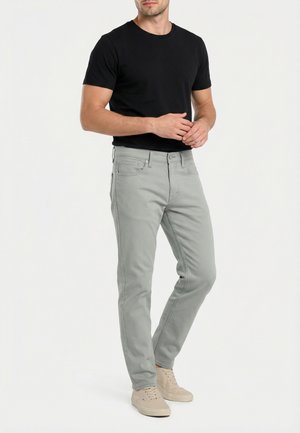 Man draagt een zwart shirt met korte mouwen, lichtgrijze spijkerbroek en beige sneakers, staand met gevouwen handen tegen een effen witte achtergrond.