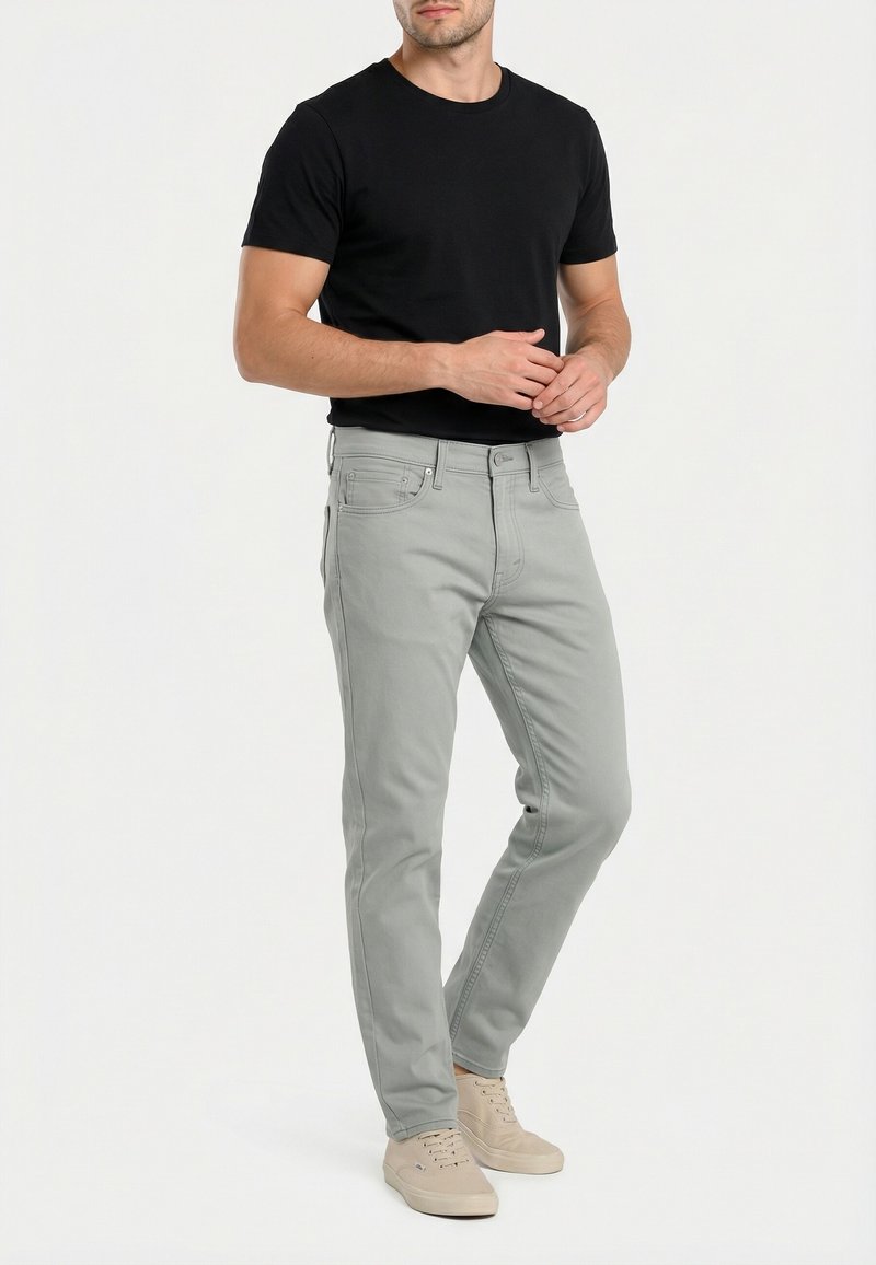 Hombre con camisa negra de manga corta, vaqueros gris claro y zapatillas beige, de pie con las manos juntas sobre un fondo blanco liso.