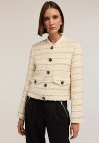 Lyhyt, kerma värinen tweed-takki, jossa on tekstuuri pintana, mustat napit ja kontrastiväri kaulus. Sisältää kaksi etutaskua.