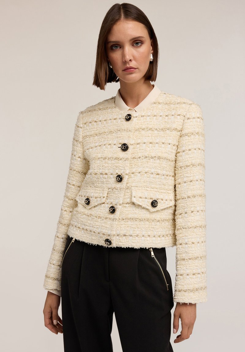 Lyhyt, kerma värinen tweed-takki, jossa on tekstuuri pintana, mustat napit ja kontrastiväri kaulus. Sisältää kaksi etutaskua.