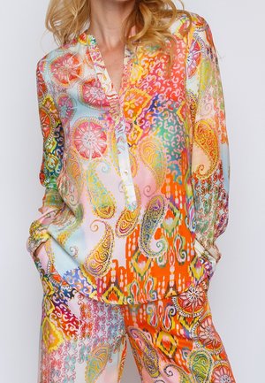 Frau trägt eine bunte Bluse mit Paisley- und Blumenmuster, langärmlig, und passende Hose, die Hände in den Taschen, vor einfachem Hintergrund.