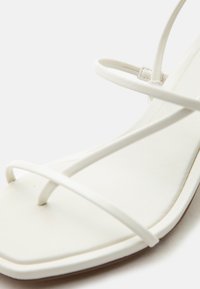 Λευκό σανδάλι με λουράκια, τετράγωνη μύτη, λεπτά λουράκια που διασχίζουν το πόδι και τον αστράγαλο, και λείο, επίπεδο πέλμα.