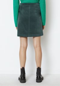 Dunkelgrüner Wildlederrock mit geradem Schnitt, ausgestattet mit zwei Gesäßtaschen und einer glatten Textur, kombiniert mit schwarzen Knöchelstiefeln.