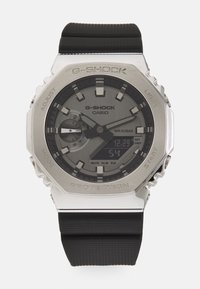 Montre Casio G-Shock avec un boîtier en acier inoxydable argenté, un cadran gris, des accents noirs, un affichage numérique et un bracelet en caoutchouc noir texturé.